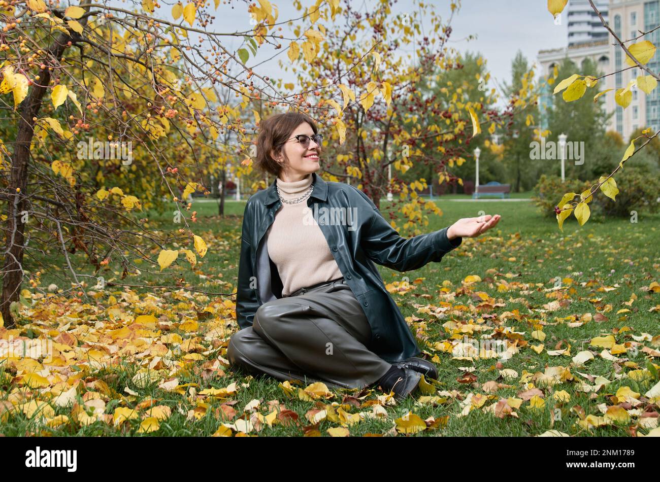 Une femme vêtue de cuir est assise sur le sol au milieu de feuilles jaunes dans un parc d'automne Banque D'Images