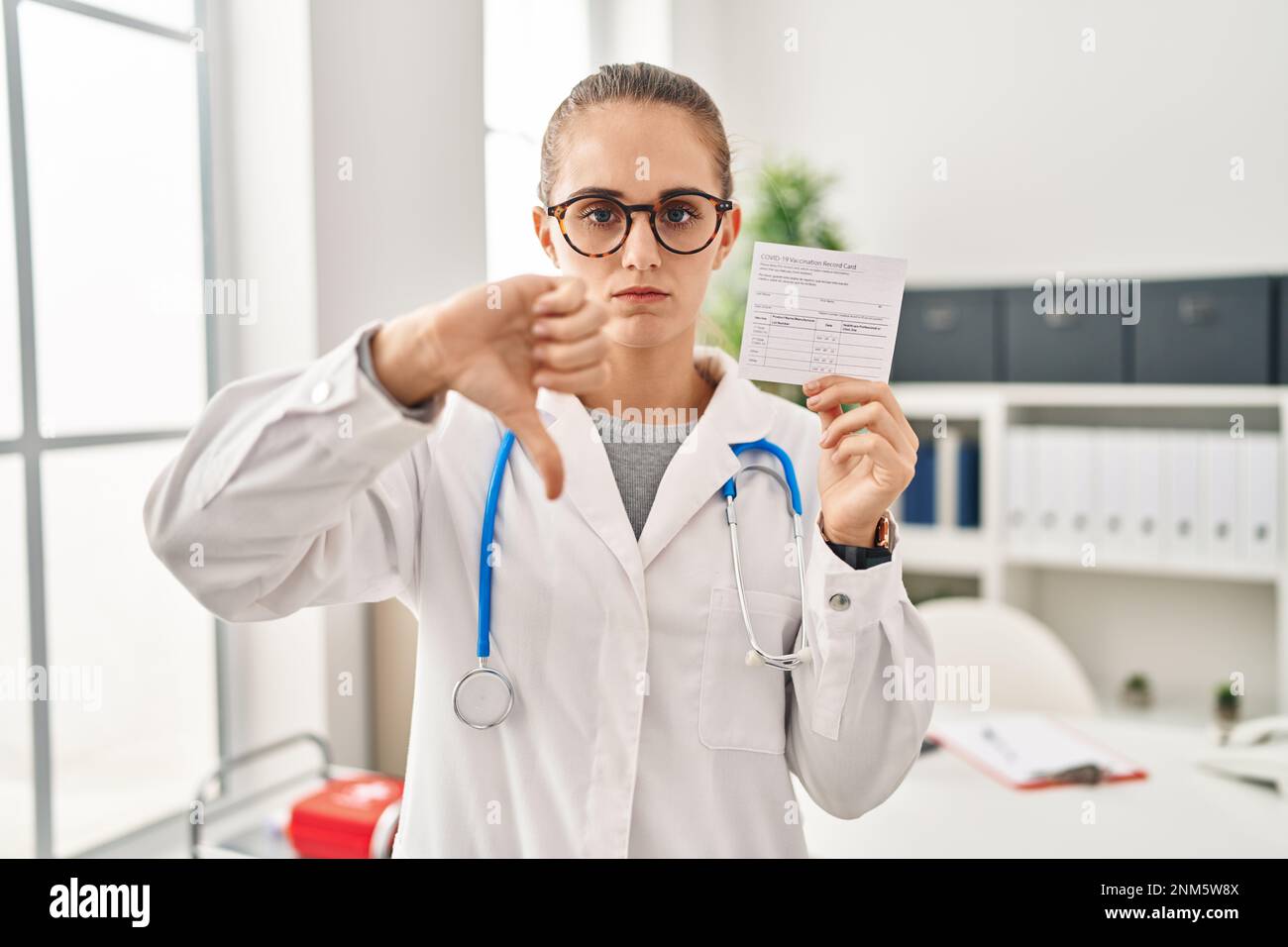 Jeune médecin femme tenant un certificat de covid avec le visage en colère, signe négatif montrant l'aversion avec les pouces vers le bas, concept de rejet Banque D'Images