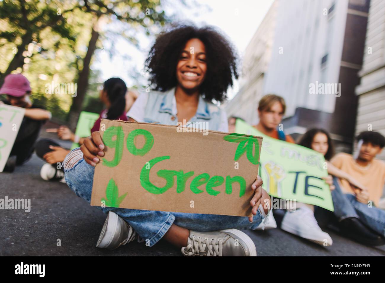 Bonne adolescente tenant une affiche « Go green » tout en étant assise avec un groupe de manifestants lors d'une manifestation contre le changement climatique. Les jeunes militants multiculturels Banque D'Images