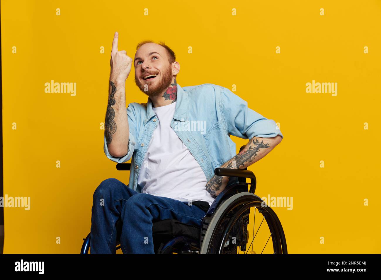Un homme en fauteuil roulant a des problèmes avec le système musculo-squelettique regarde la caméra montre un doigt dessus, avec des tatouages sur ses mains assis sur un jaune Banque D'Images