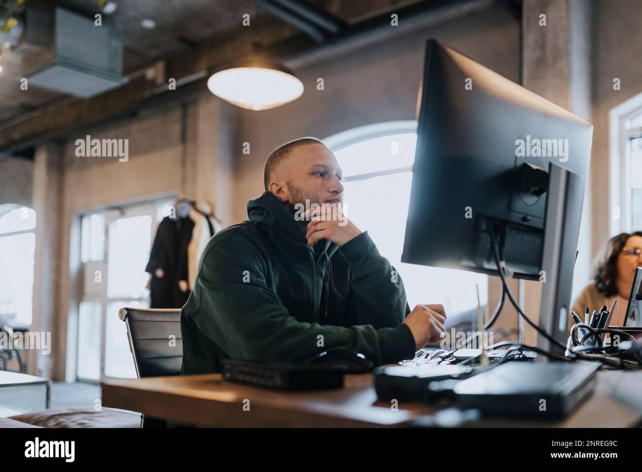 Un jeune hacker mâle avec la main sur le menton utilisant un ordinateur au bureau dans le bureau de création Banque D'Images