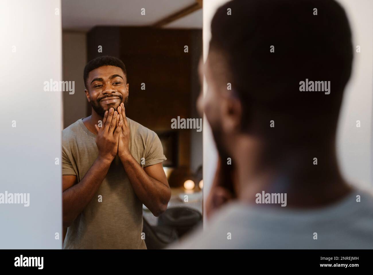 Jeune homme souriant touchant le visage tout en regardant dans le miroir de la maison Banque D'Images