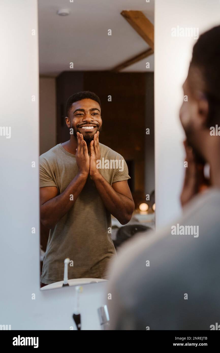 Homme souriant appliquant un hydratant tout en regardant dans le miroir à la maison Banque D'Images