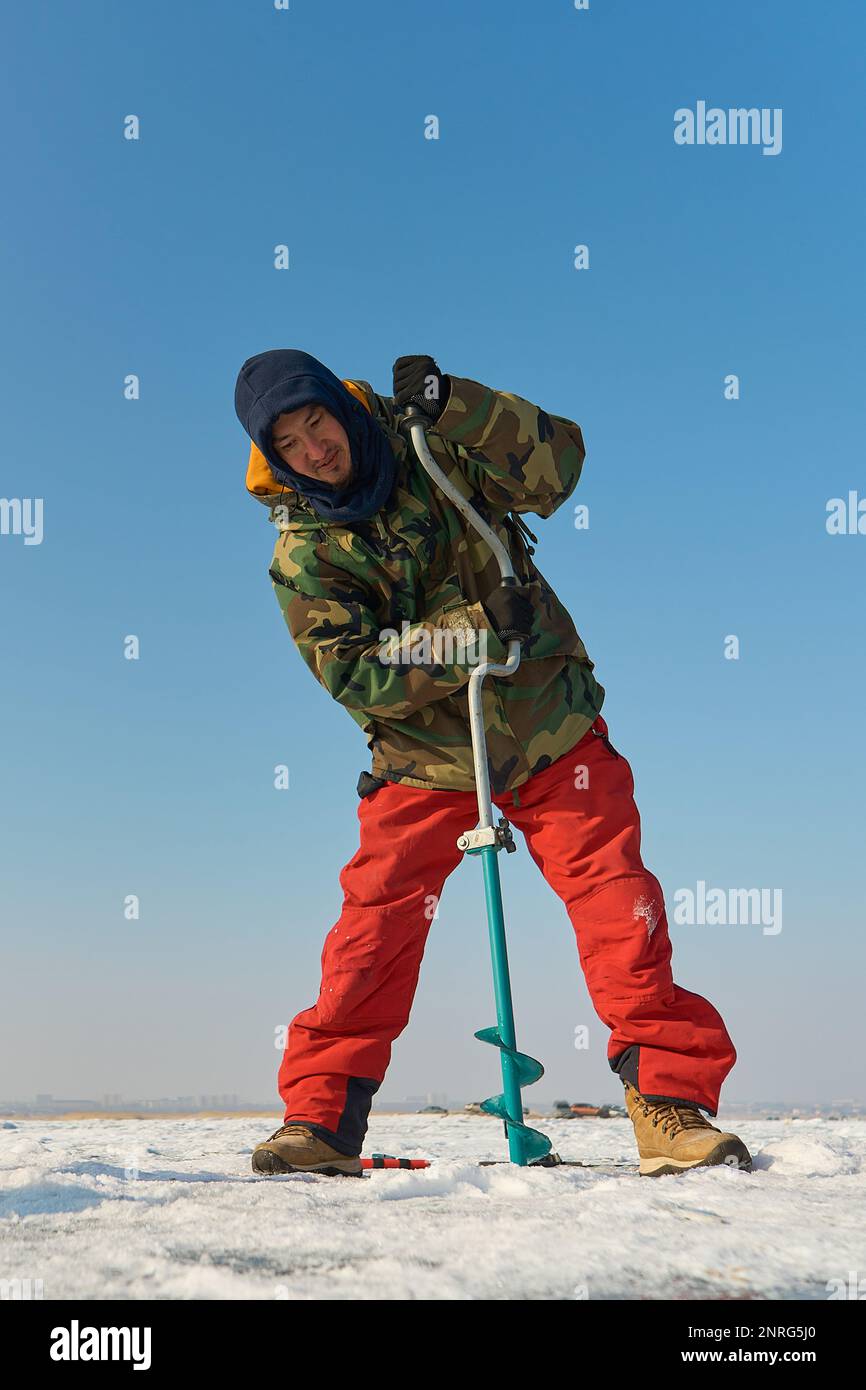 Un homme asiatique aux couleurs chaudes fore un trou dans la glace en hiver Banque D'Images