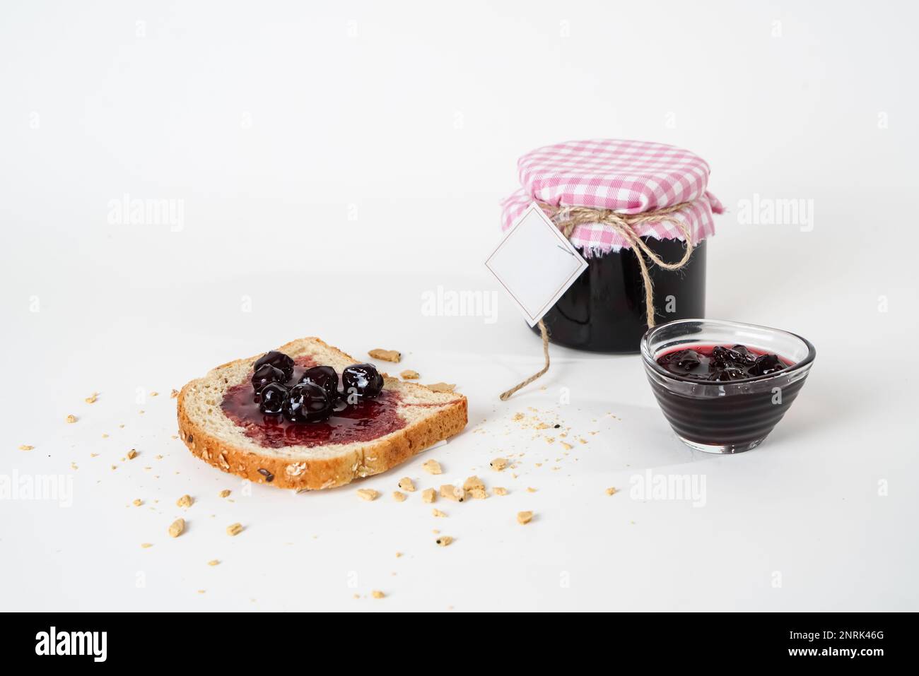 Récipient traditionnel de confiture de cerises acides avec étiquette vide, petit bol et une tranche de pain avec confiture isolée sur fond blanc. Banque D'Images