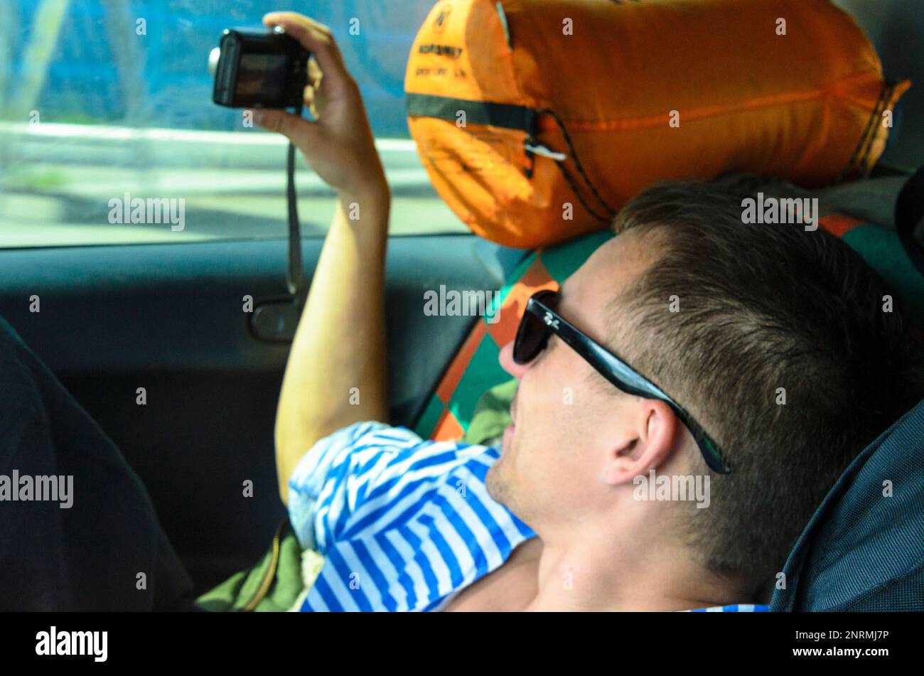 Un homme qui se repose dans des lunettes sombres est photographié à travers le verre d'une voiture dans le siège arrière parmi les choses touristiques. Banque D'Images