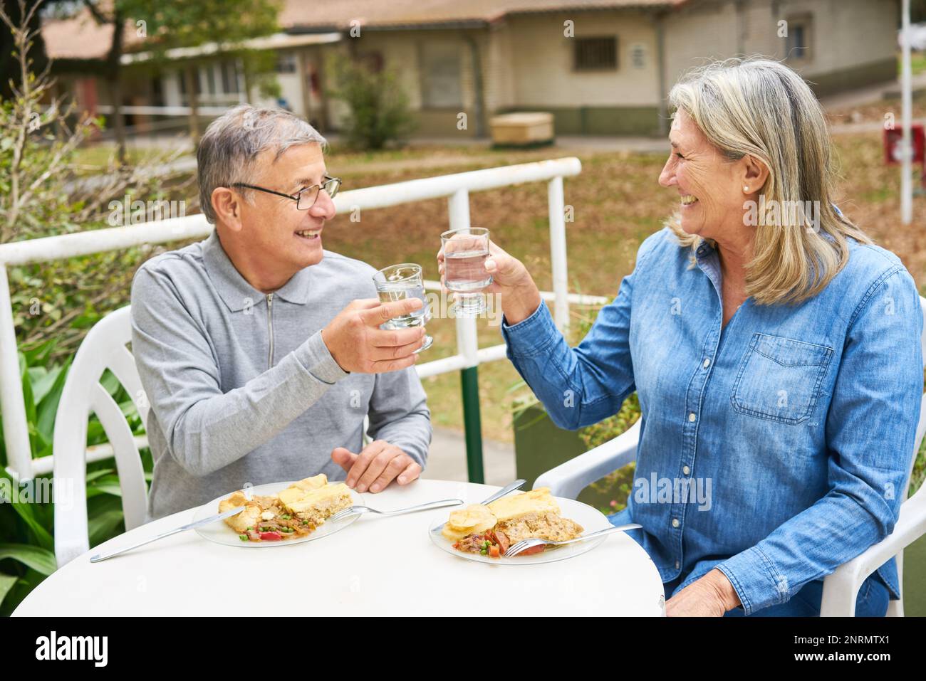 Homme et femme joyeux et senior toaster un verre d'eau tout en prenant un repas dans le jardin de la maison de retraite Banque D'Images