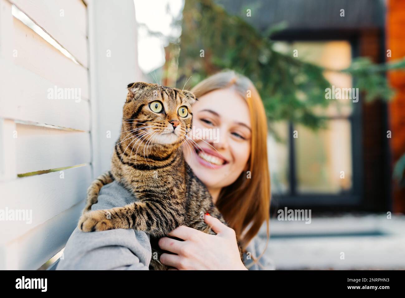 Jolie jeune femme blonde souriante tient un chat écossais aux yeux verts dans ses bras et l'épouse à l'extérieur, concept d'aimer et de s'occuper des animaux de compagnie Banque D'Images