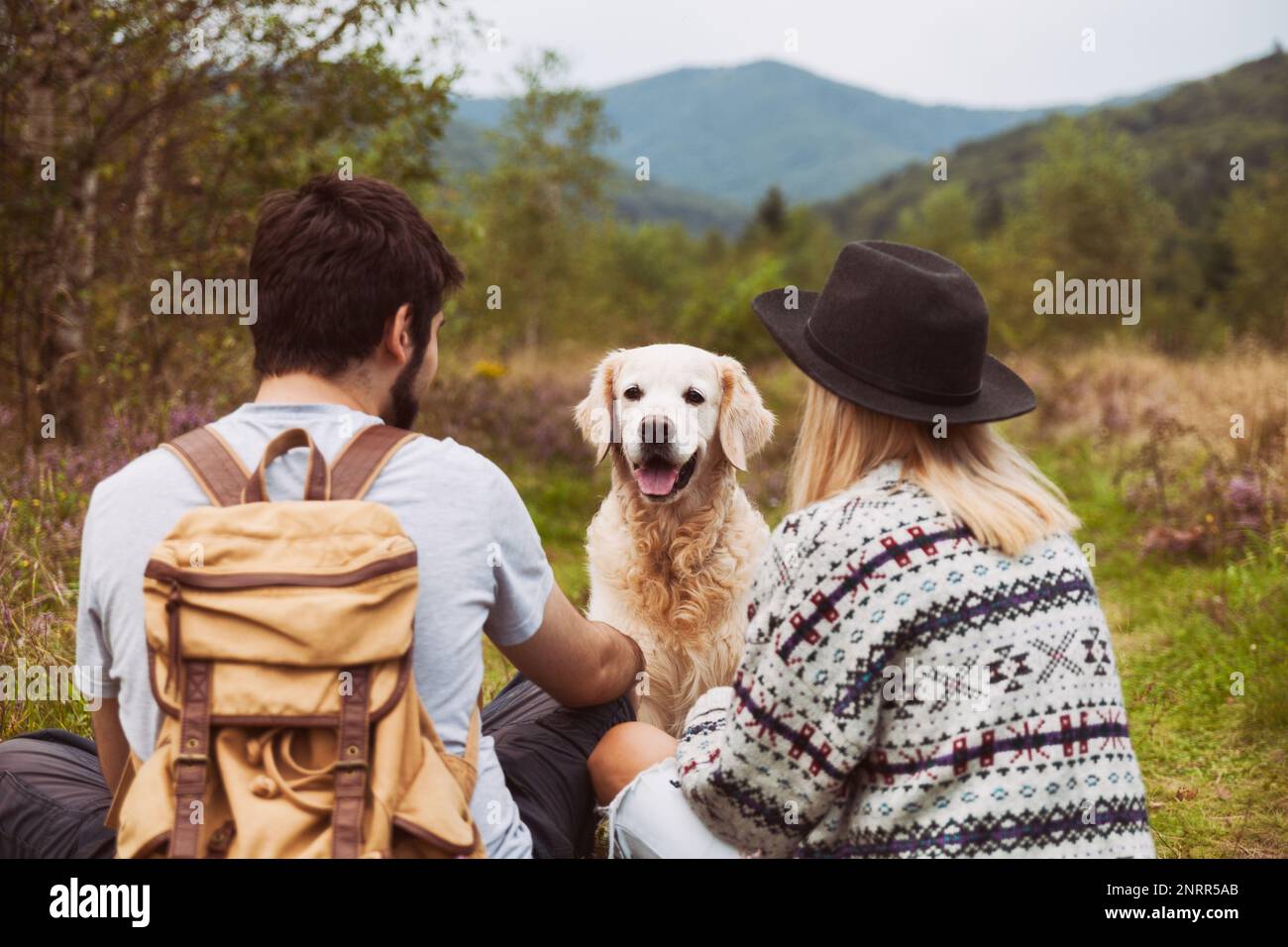 Jeune couple avec un heureux chien Golden Retriever souriant assis sur l'herbe dans la vallée des montagnes d'été. Animaux de compagnie Voyage, randonnée et activités de week-end concept. Banque D'Images