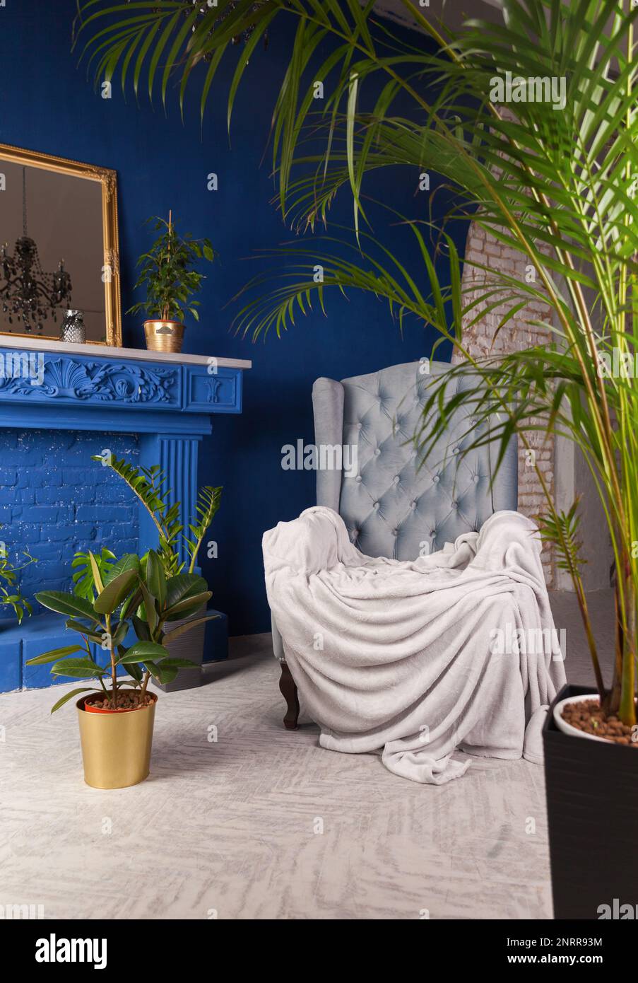 Manteau gris sur le fauteuil près du mur bleu et cheminée décorative dans la maison ou l'hôtel de charme. Plantes vertes dans le salon intérieur dans l'appartement art déco. Banque D'Images
