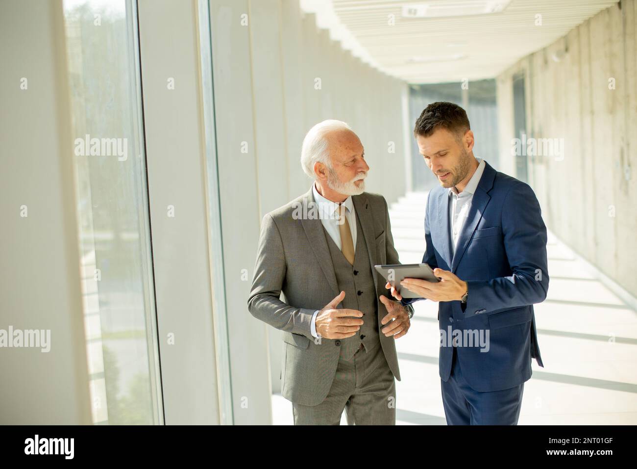 Beau homme d'affaires mature et son jeune collègue discutant du rapport final sur les tablettes numériques dans le couloir du bureau Banque D'Images