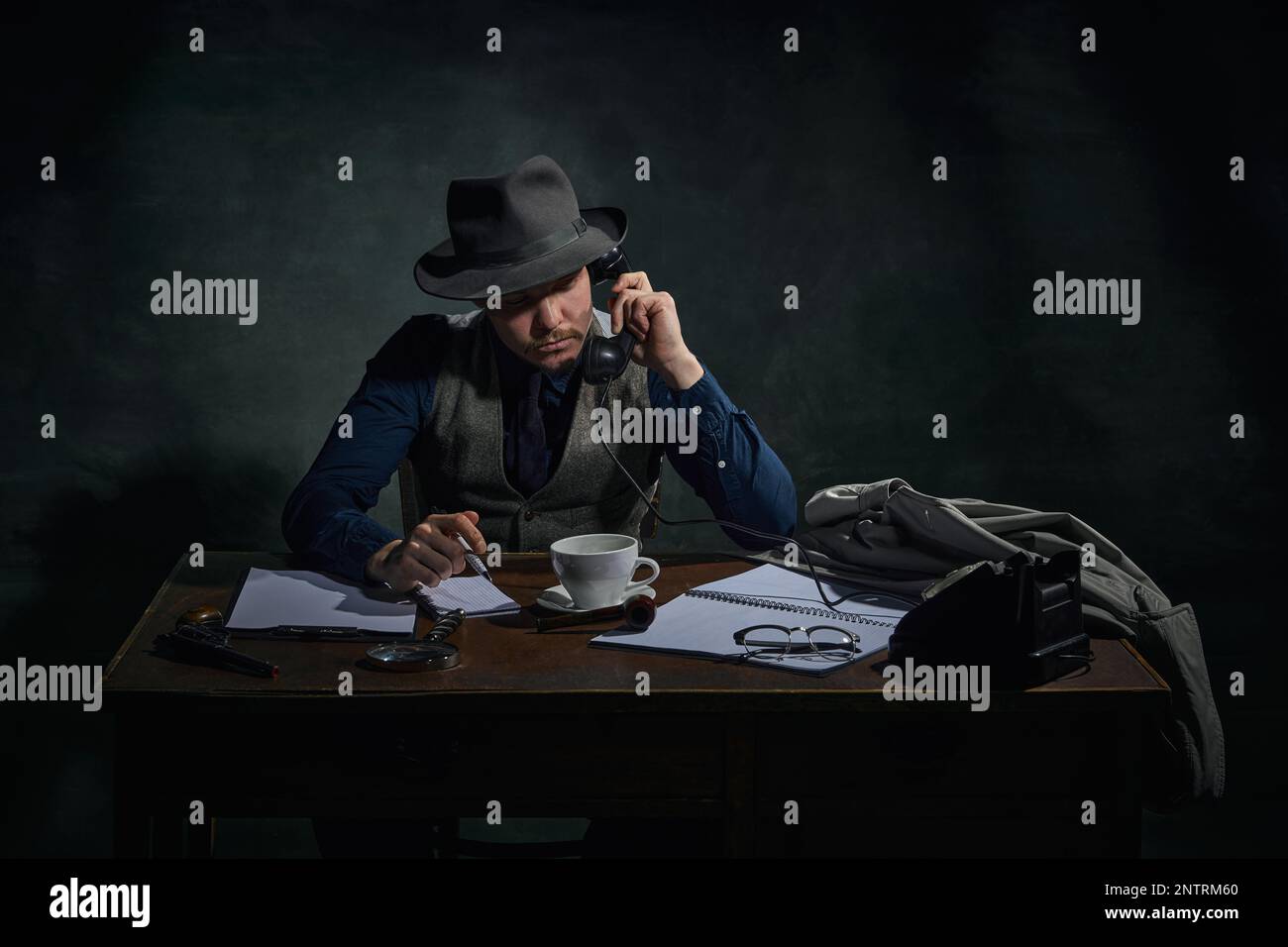 Détective professionnel en chapeau fedora assis à table et parlant au téléphone sur fond vert foncé vintage. Enquête Banque D'Images