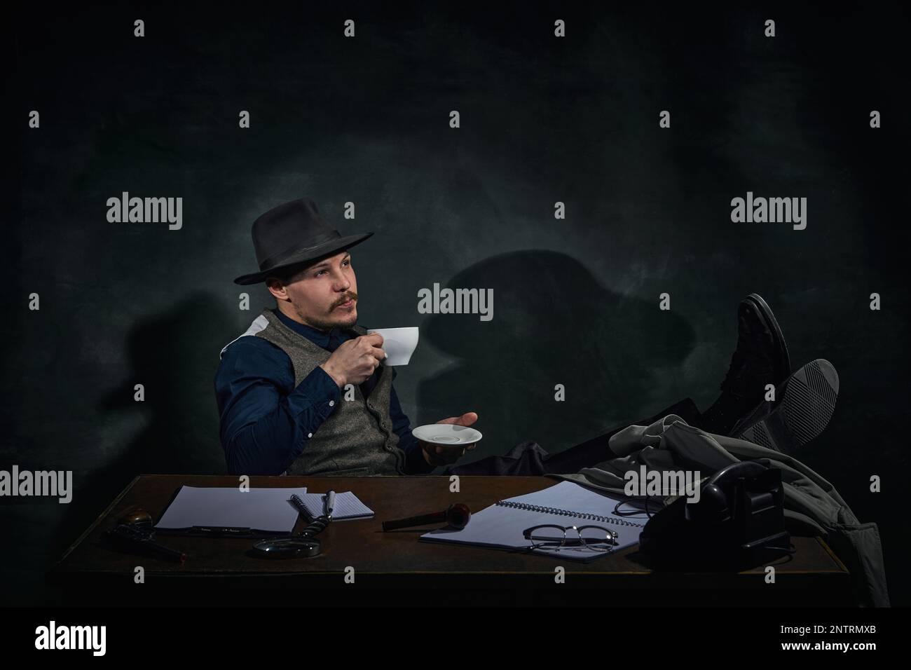 Détective professionnel dans le chapeau fedora assis à table avec le visage réfléchi et boire un café sur fond vert foncé vintage. Inspecteur Banque D'Images