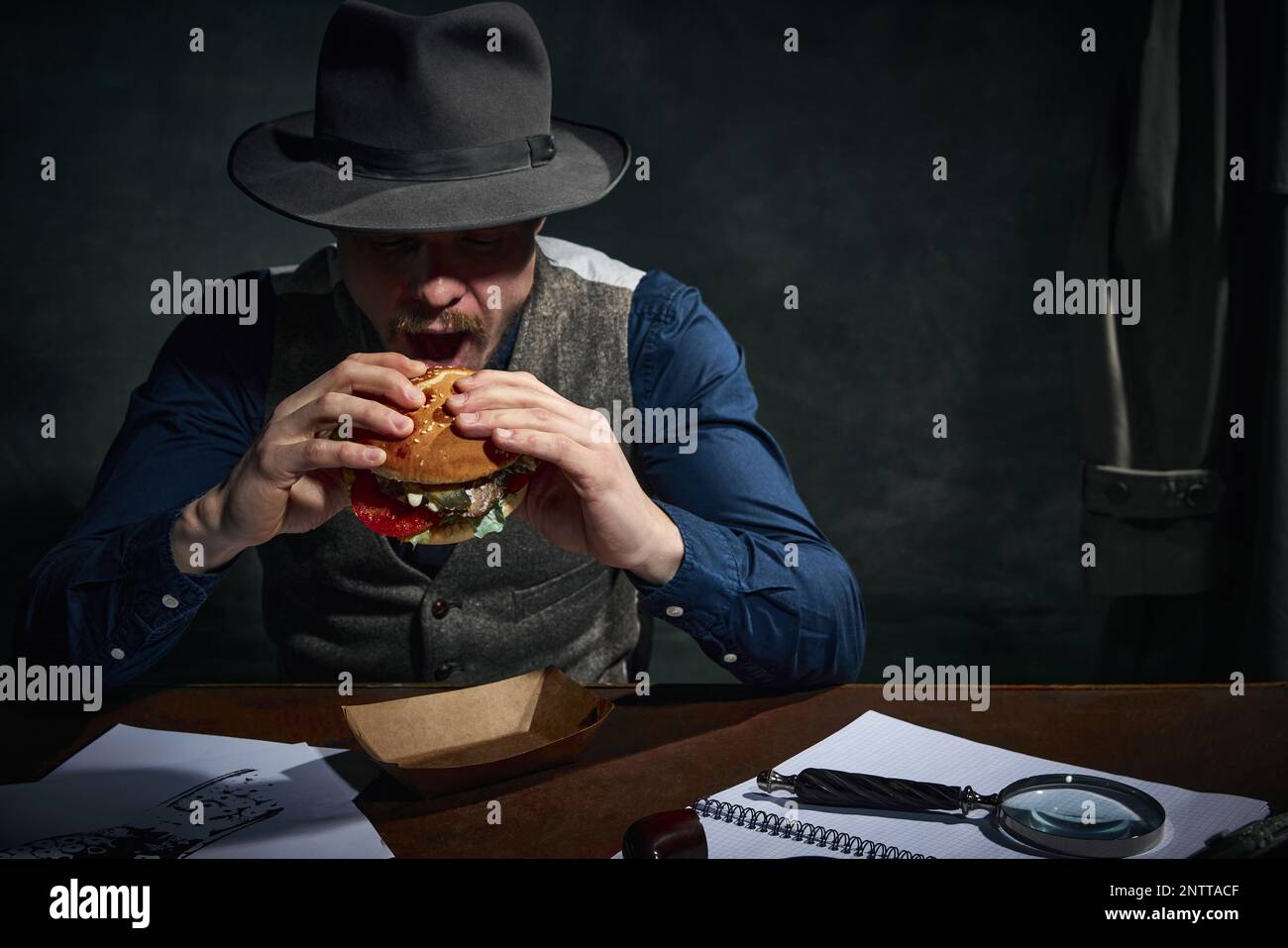 Détective professionnel dans le chapeau fedora assis à table et manger délicieux hamburger pour le déjeuner sur fond vert foncé vintage Banque D'Images