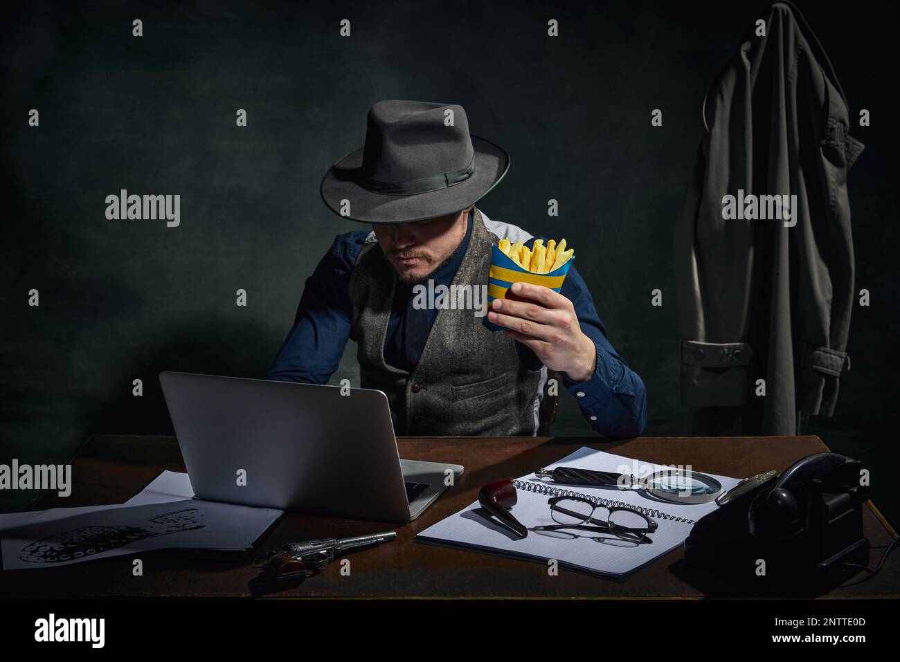 Détective professionnel en chapeau fedora assis à table, travaillant sur ordinateur portable et mangeant des frites sur fond vert foncé vintage Banque D'Images