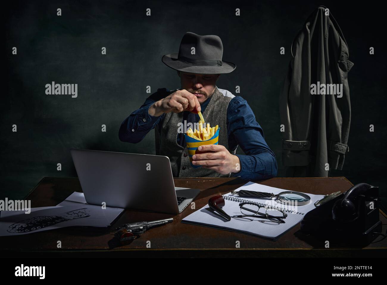 Détective professionnel en chapeau fedora assis à table, travaillant sur ordinateur portable et mangeant des frites sur fond vert foncé vintage. Homme occupé Banque D'Images