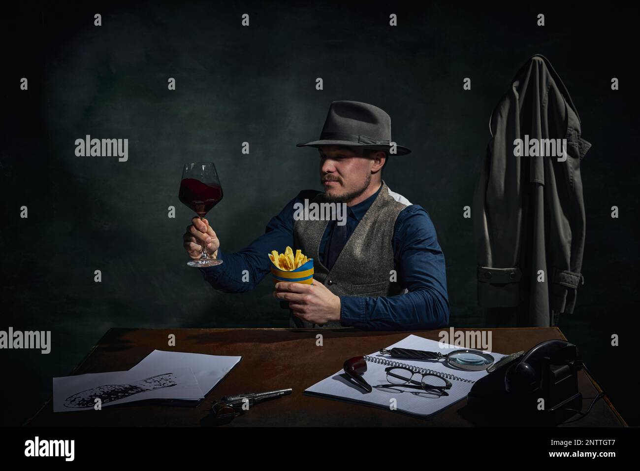 Détective professionnel dans le chapeau fedora assis à table, boire du vin rouge et manger des frites sur fond vert foncé vintage Banque D'Images