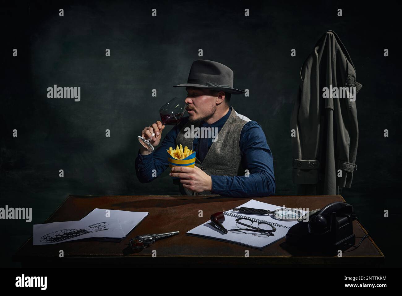 Détective professionnel dans le chapeau fedora assis à table, boire du vin rouge et manger des frites sur fond vert foncé vintage Banque D'Images