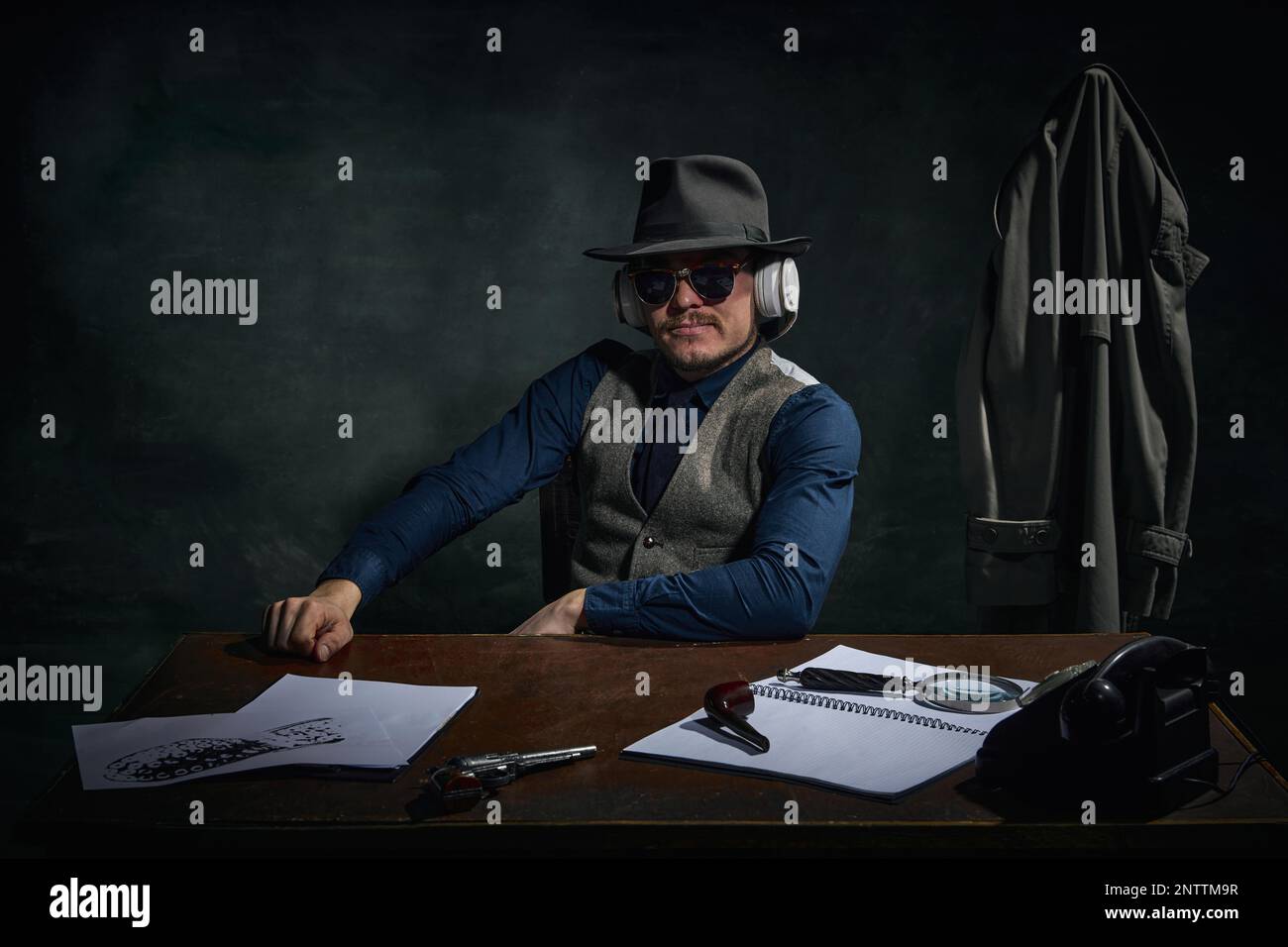 Homme élégant, détective professionnel en chapeau fedora et lunettes de soleil assis à la table et écouter des informations dans les écouteurs sur le vert foncé vintage Banque D'Images