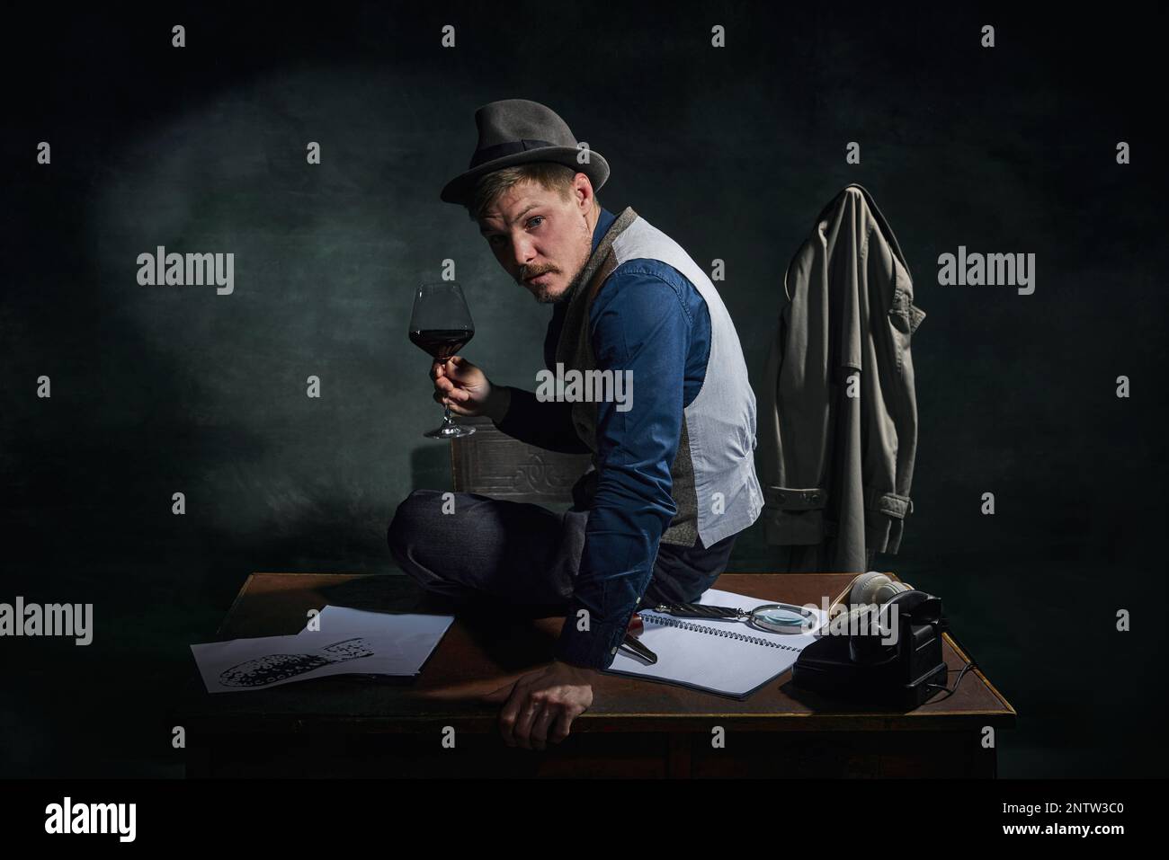 Détective professionnel en chapeau fedora et vêtements classiques assis sur une table avec un verre de vin rouge sur fond vert foncé vintage Banque D'Images