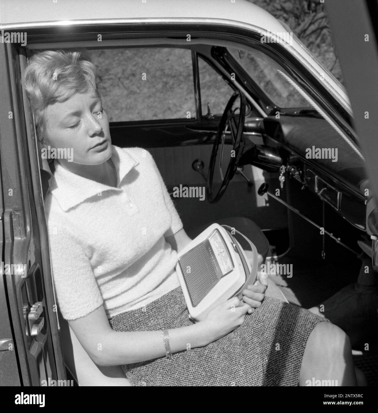 Dans le 1960s. Une jeune femme dans une voiture qui écoute une radio portable. Radio à transistor à piles avec bandes AM et FM. Les stations de radio et les fréquences sont affichées à l'avant et un bouton permet des tourner et des régler pour obtenir la meilleure réception possible. Suède 1960 Conard réf. 4220 Banque D'Images