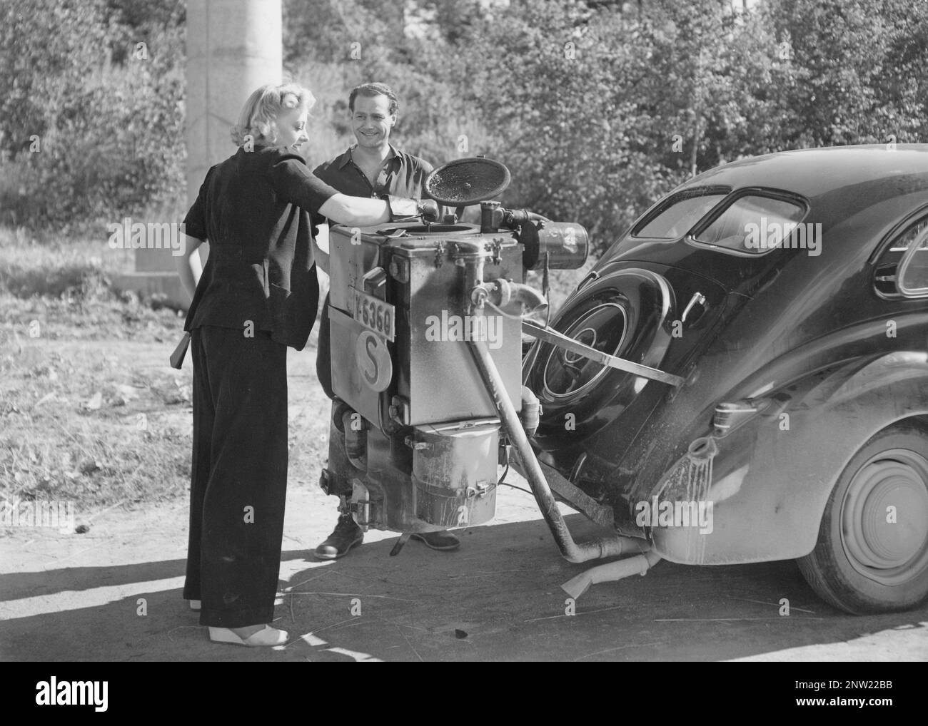Le gaz de bois dans les années 1940 L'enregistrement du long métrage suédois Stora skrällen de 1942 où les acteurs Karin Ekelund et Allan Bohlin participent. Une caractéristique naturelle pendant ces années pendant la seconde Guerre mondiale est l'unité de gazéification du bois sur la voiture. Le gaz de bois est un gaz qui résulte de la combustion incomplète du bois ou du charbon, et qui a été utilisé pour alimenter les véhicules. Le conducteur devait remplir lui-même le charbon et s'assurer que la combustion fonctionnait comme il se devait. En raison d'un manque de carburant pendant la seconde Guerre mondiale, le gaz de bois a joué un rôle majeur dans le maintien des transports. Banque D'Images