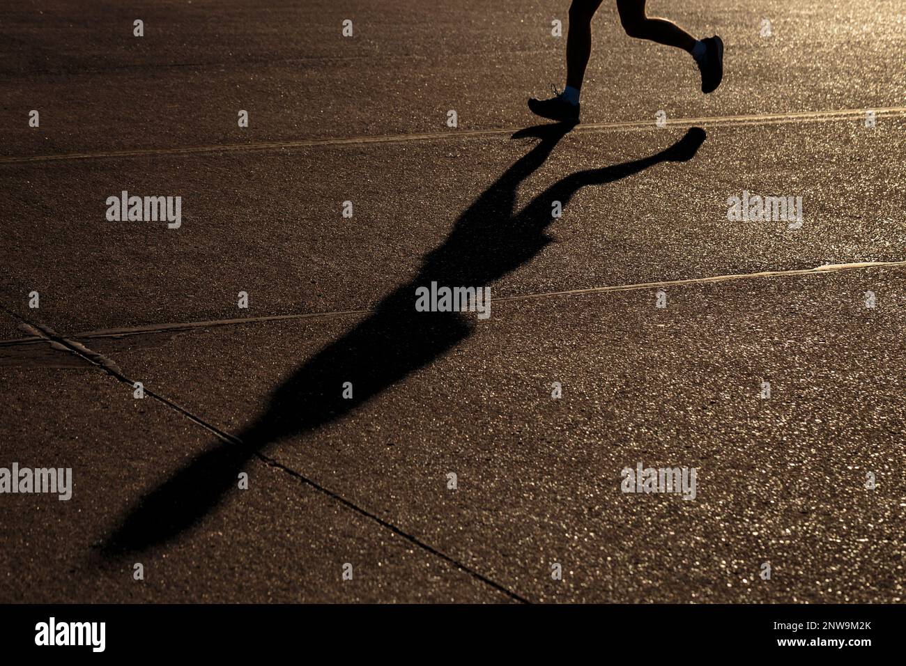 Jambes d'un jogging masculin simple méconnaissable qui court dans la ville au coucher du soleil, avec sa longue ombre projetée sur un trottoir sombre Banque D'Images