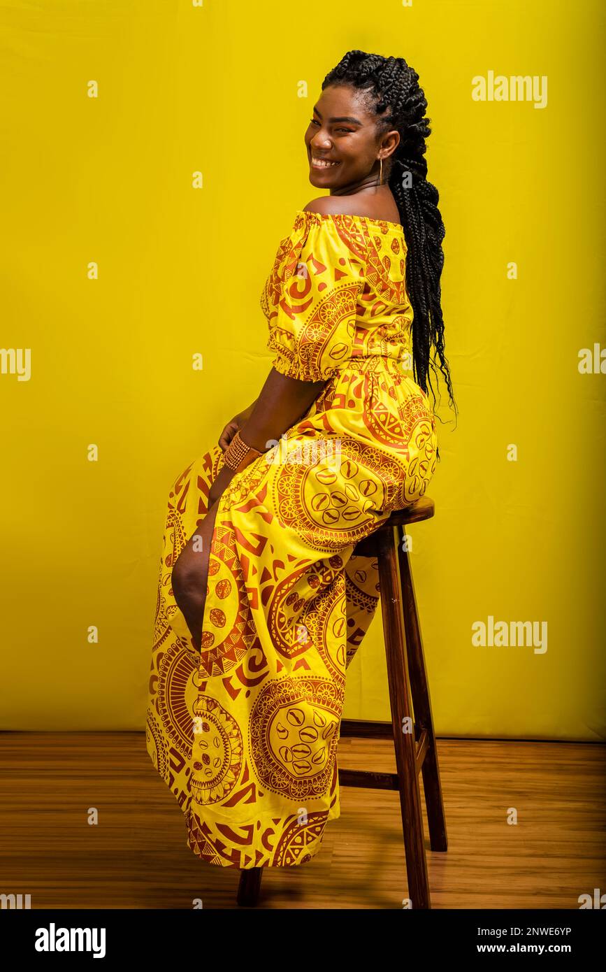 Belle jeune femme assise sur un tabouret. Vêtements colorés. Isolé sur fond jaune. Banque D'Images