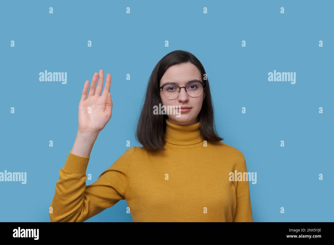 Une jeune fille étudiante avec des lunettes et un chandail jaune chaud agitant sa salutation à la main ou en disant au revoir sur un fond bleu. Communication non verbale Banque D'Images