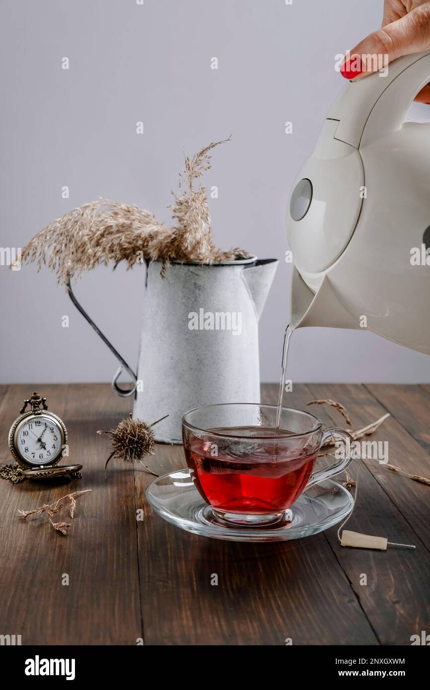Une main femelle verse de l'eau chaude d'une bouilloire dans une tasse en verre transparent pour faire du thé, avec une horloge vintage et un vase de plantes sèches à côté Banque D'Images
