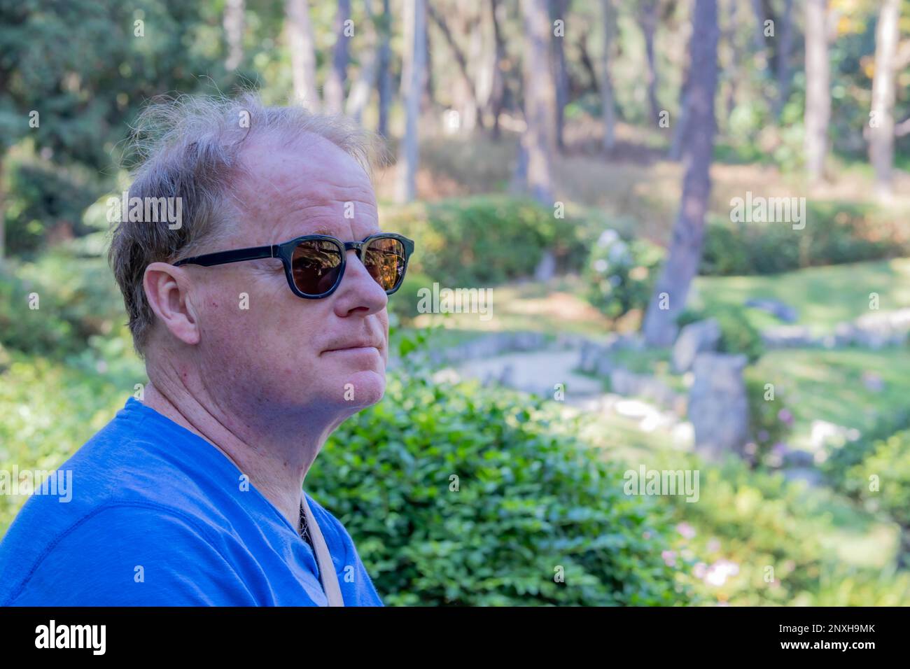 Beau et pensif hollandais senior homme dans des lunettes de soleil, profil regardant droit devant, portant blouse bleue, étang, buissons verts et arbres dans le dos flou Banque D'Images