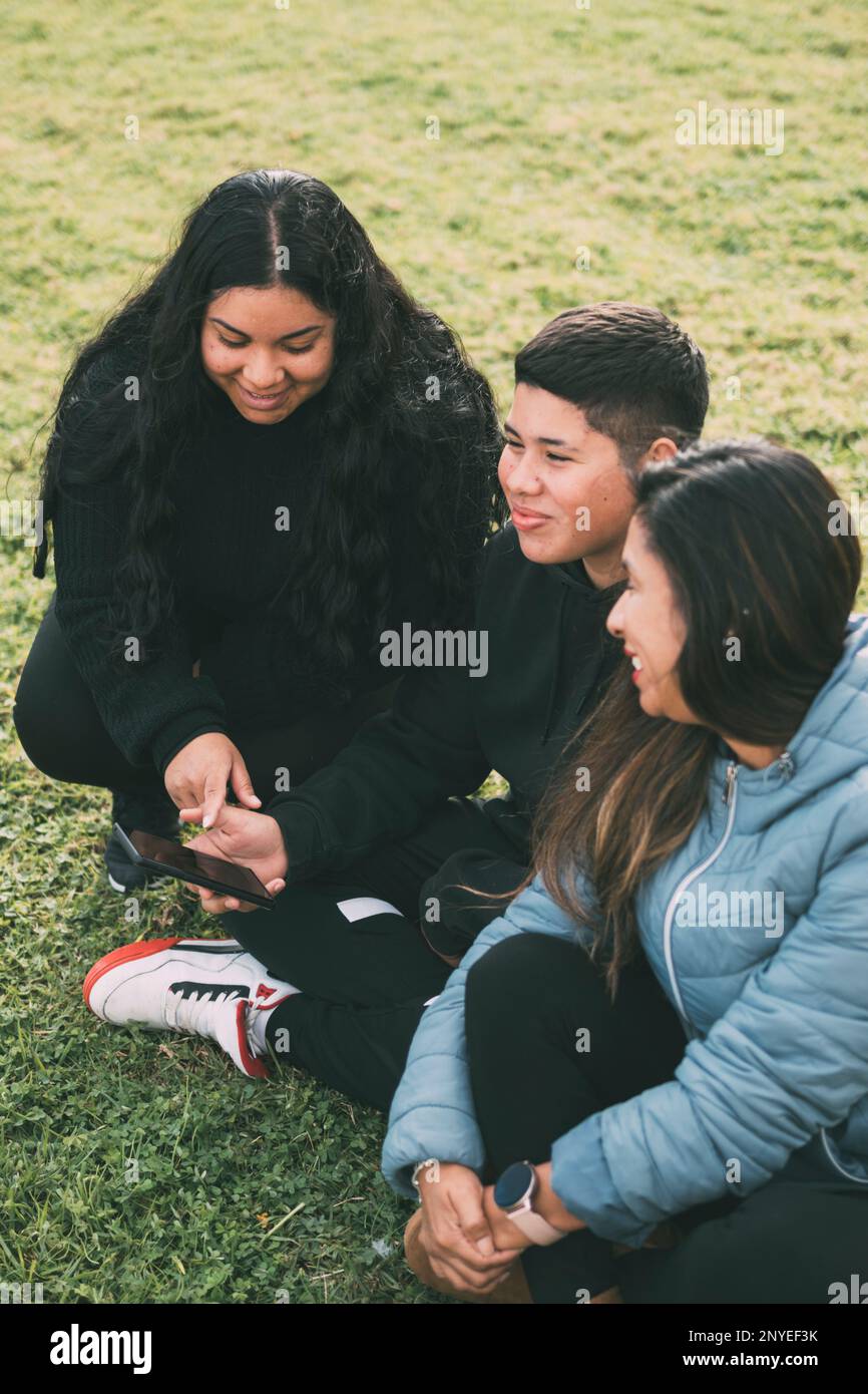 Trois individus d'origine hispanique-latino, assis sur l'herbe verte luxuriante d'un parc, avec un appareil intelligent. Des expressions relaxantes et souriantes sur leur Banque D'Images