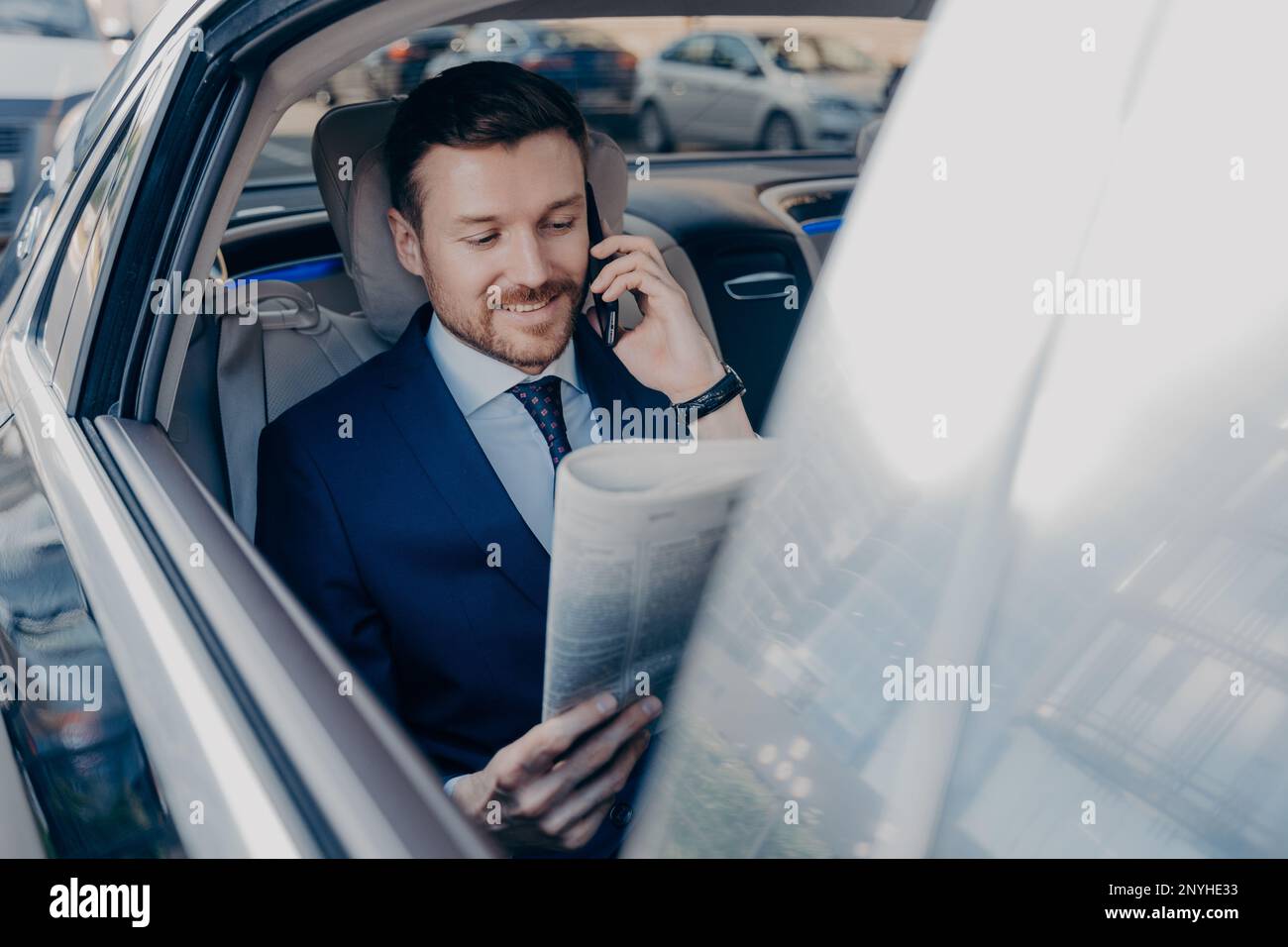Bon look Happy Executive Manager en tenue élégante et formelle lit le journal sur le siège arrière de la voiture en même temps parlant sur smartphone avec la partie d'affaires Banque D'Images