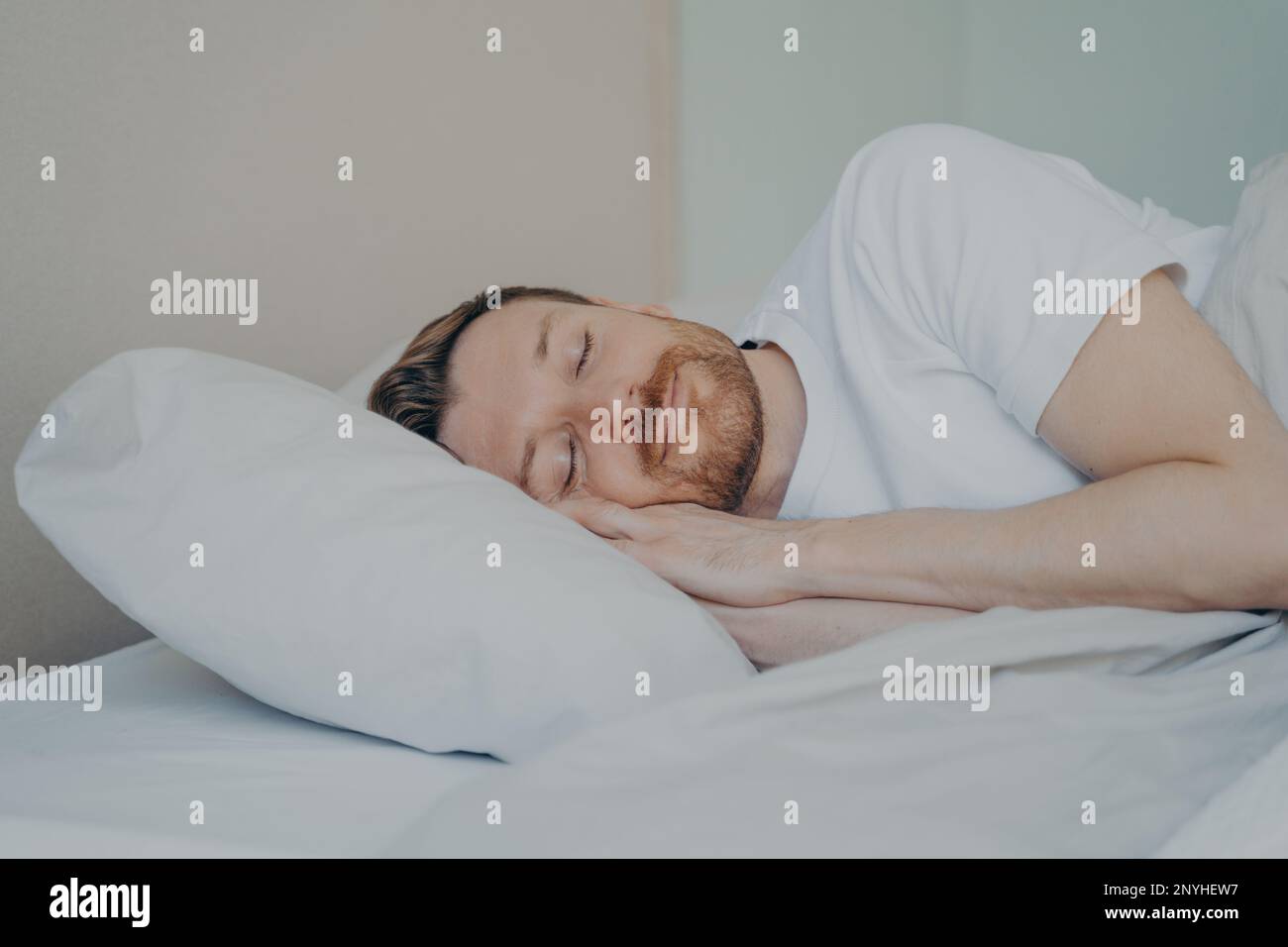 Gros plan photo d'un jeune homme barbu avec les yeux fermés s'endormir pendant ses vacances le dimanche, dormant avec le sourire tout en étant allongé sur un oreiller blanc en b Banque D'Images