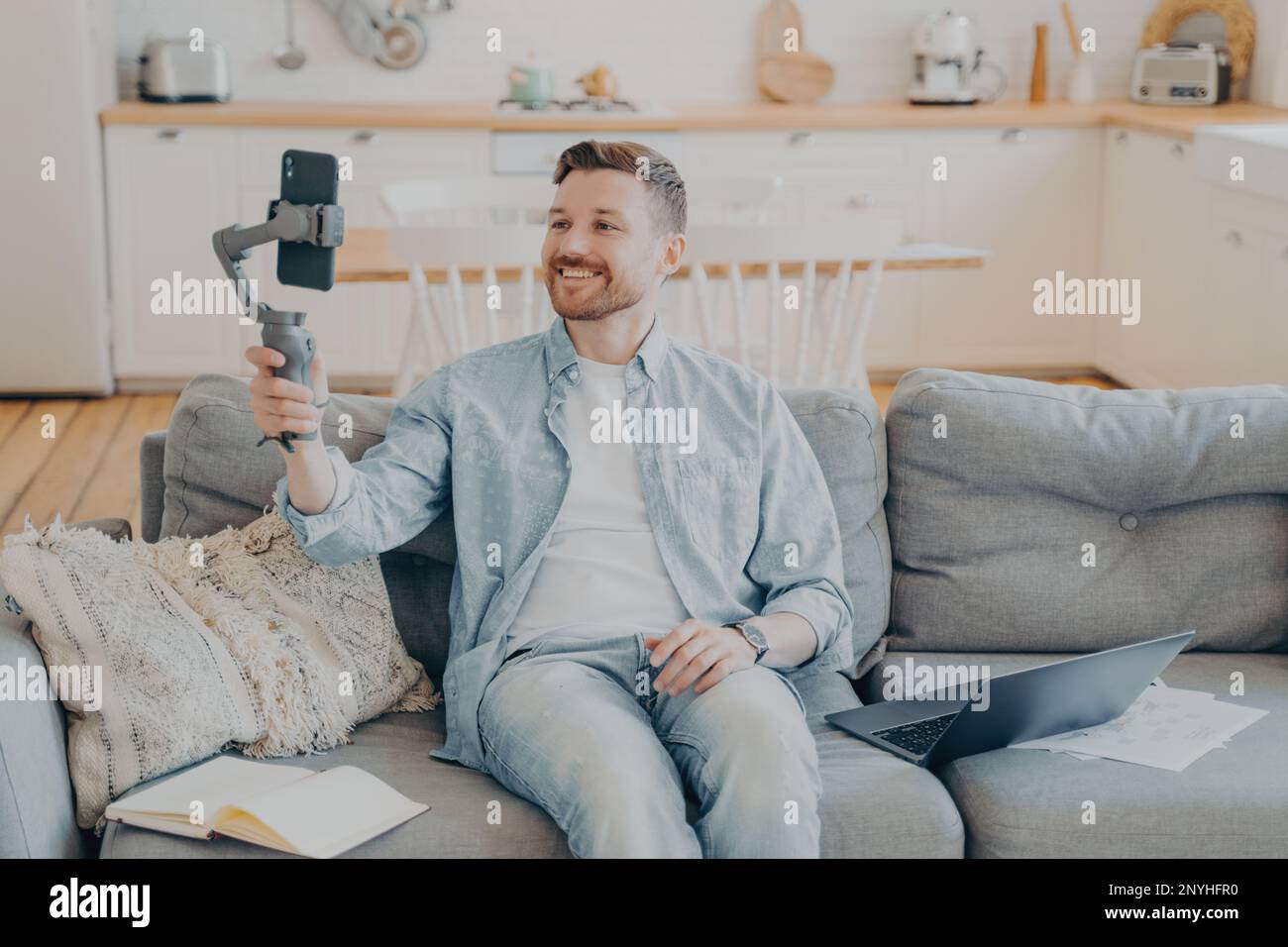 Jeune homme souriant parlant à son ami et lui montrant son appartement pendant un appel vidéo, tenant une nacelle avec un téléphone, assis sur un canapé à côté d'un ordinateur portable, Banque D'Images
