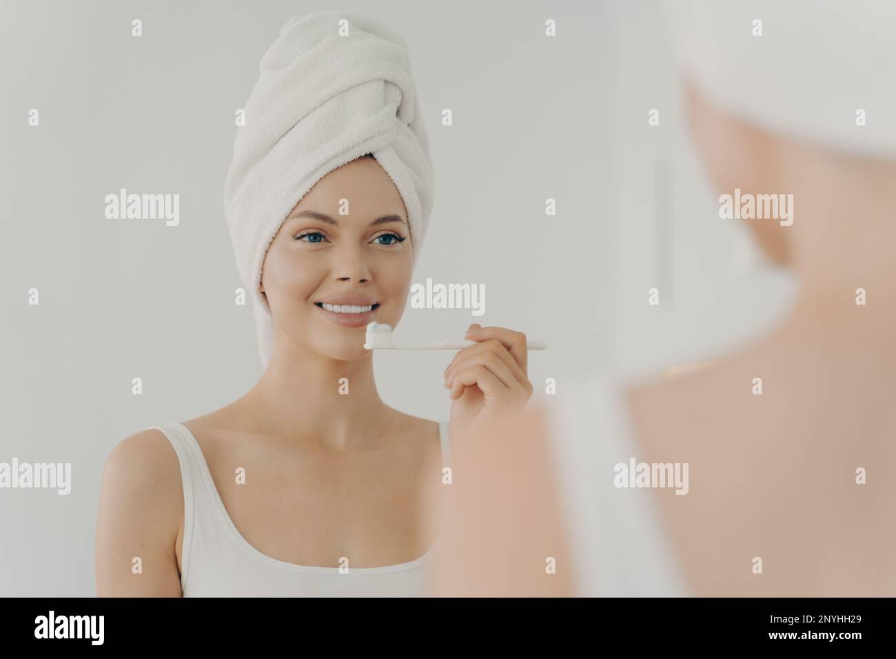 Portrait de jeune femme en bonne santé belle avec une serviette sur la tête après la douche ayant le sourire en dents de brosse doucement, regardant dans le miroir avec Banque D'Images