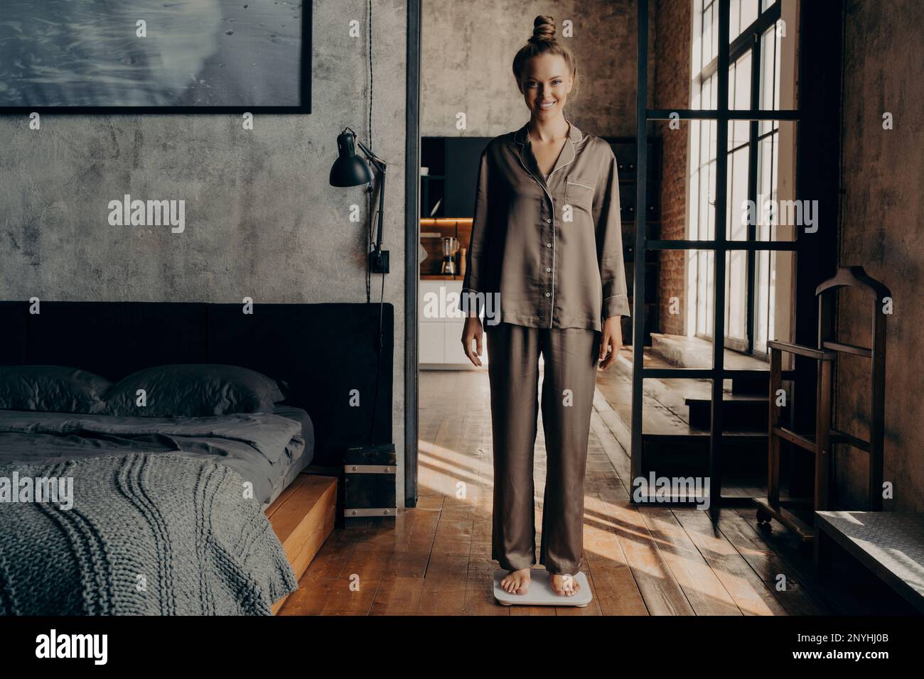 Régime et perte de poids. Pleine longueur de jeune fille attrayante heureuse avec les cheveux attachés dans un petit pain dans un pyjama en satin décontracté debout sur des balances dans un lit élégant Banque D'Images