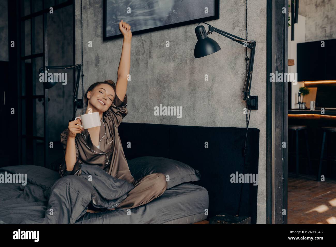 Jeune belle femme avec les yeux fermés et les cheveux attachés dans un petit pain commençant le matin avec une tasse de café chaud, s'étirant dans le lit à la maison tout en étant assis dans le siège Banque D'Images