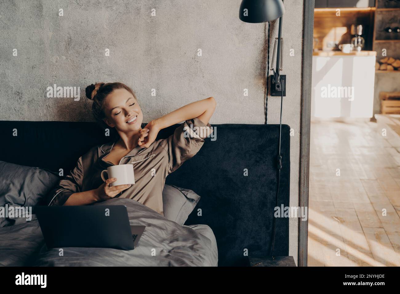 Jolie femme qui garde les yeux fermés dans un pyjama en satin tenant une tasse de café et prenant le soleil du matin, allongé dans le lit avec un ordinateur portable après s'être réveillé Banque D'Images