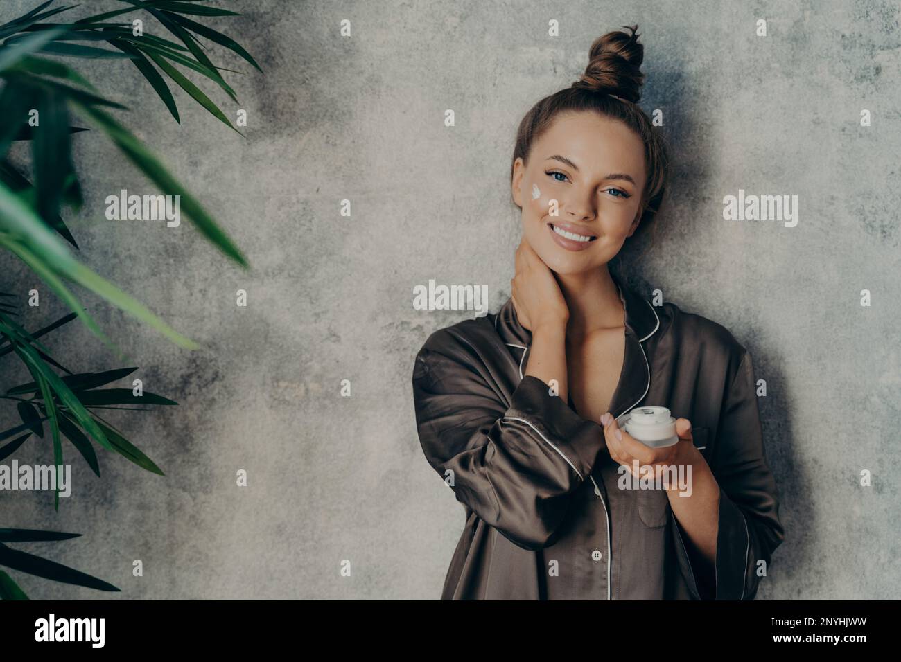 Image de femme gaie en pyjama en soie souriant tout en appliquant la crème du visage, posant avec un pot de lotion hydratante dans sa main, isolé sur béton W Banque D'Images