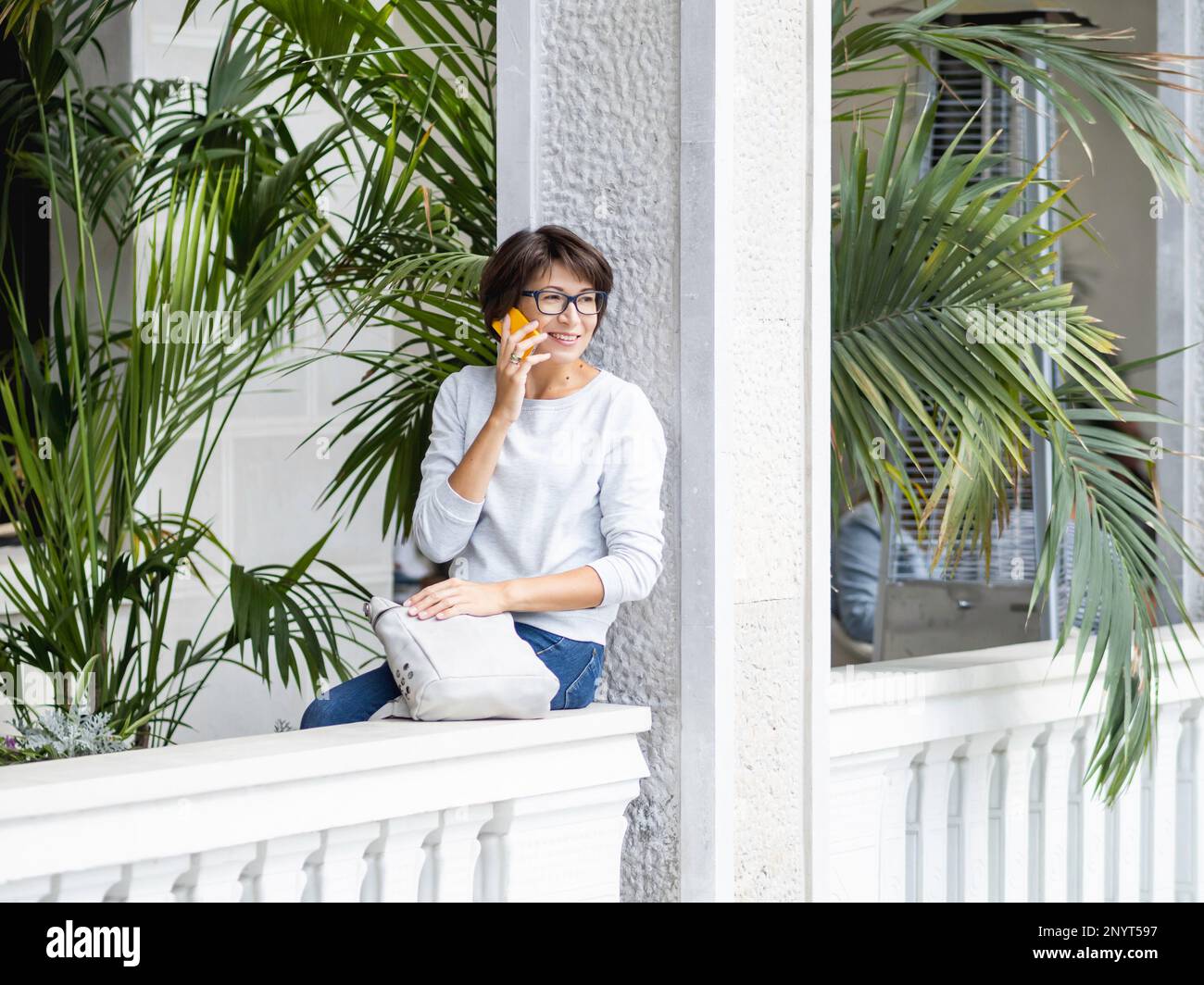 Une femme parle sur un smartphone. Jolie femme à lunettes sur un balcon avec palmiers dans des pots de fleurs. Maison tropicale. Des vibes d'été. Banque D'Images