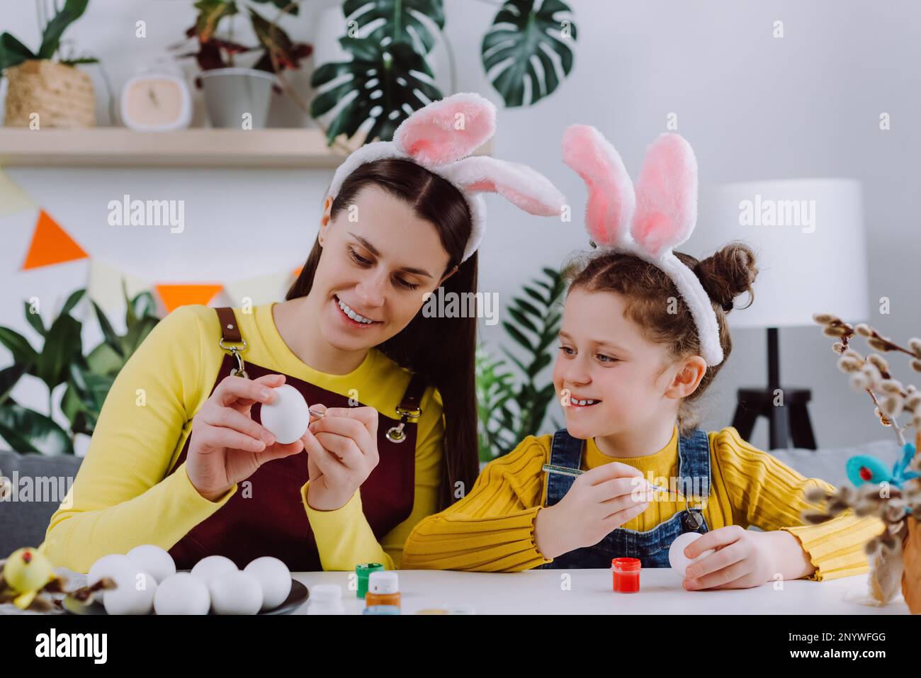 Jeune mère souriante enseignant jolie petite fille enfant peinture des œufs assis ensemble à la table, porte des oreilles de lapin. Fête de Pâques et tradition Banque D'Images