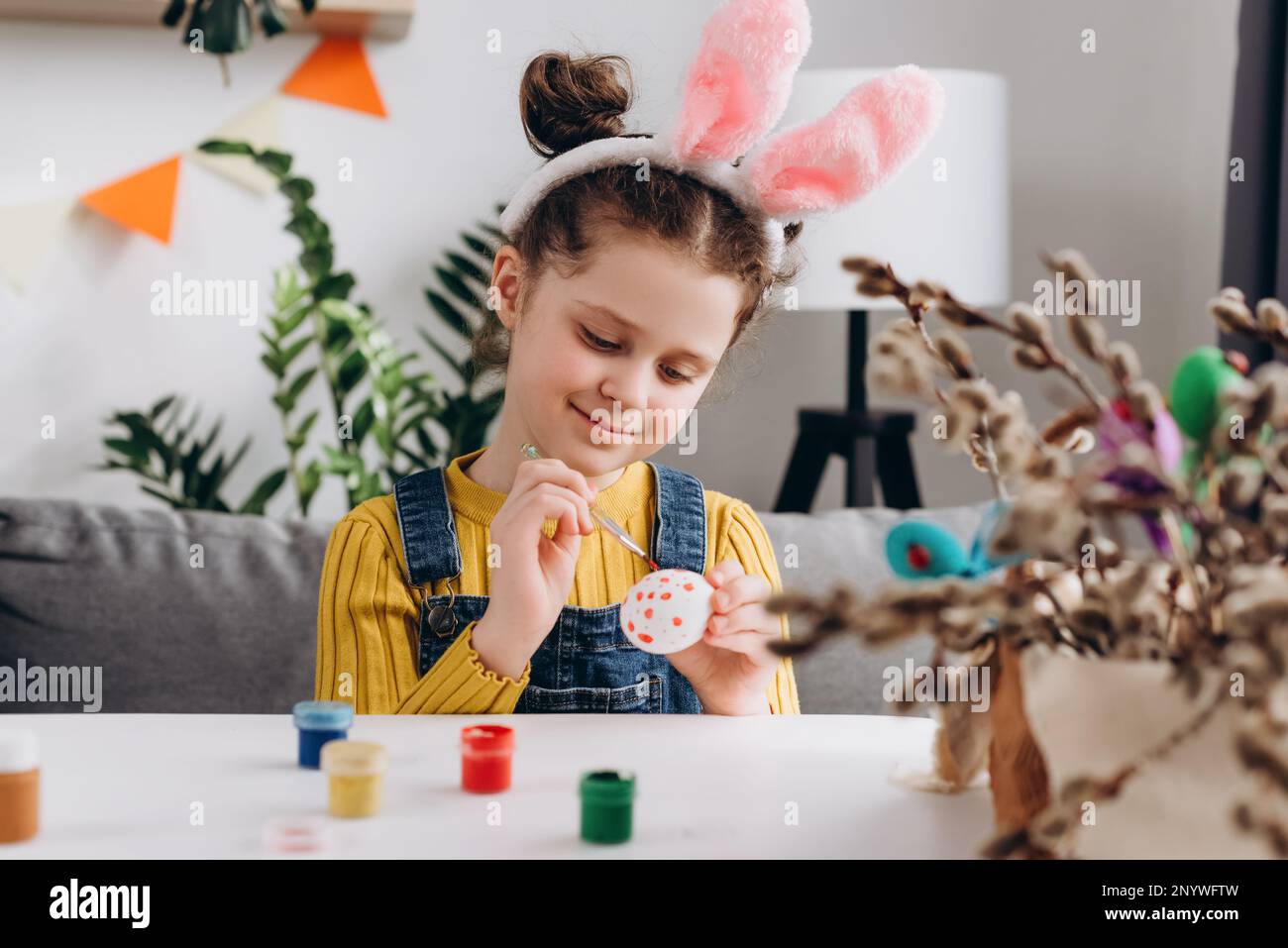 Gros plan portrait d'une adorable petite fille souriante portant des oreilles de lapin moelleuses roses assis seul à une table blanche et peignant des œufs tout en préparant FO Banque D'Images