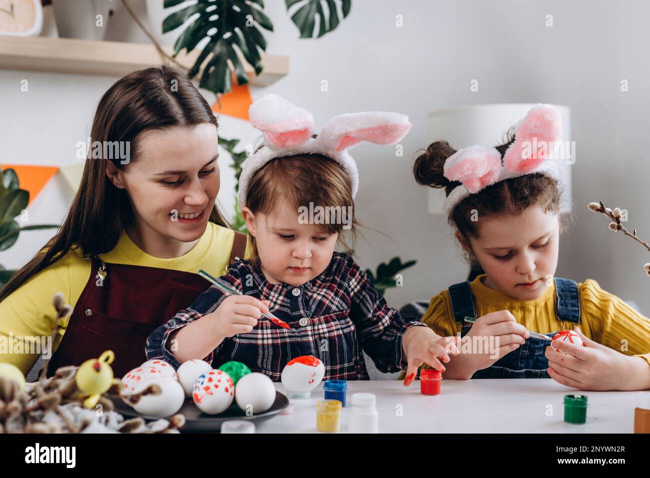 Bonne jeune maman caucasienne et deux petites filles décorant des œufs avec des peintures pour les vacances de Pâques tout en étant assis ensemble à la table blanche. Famille amusante Banque D'Images