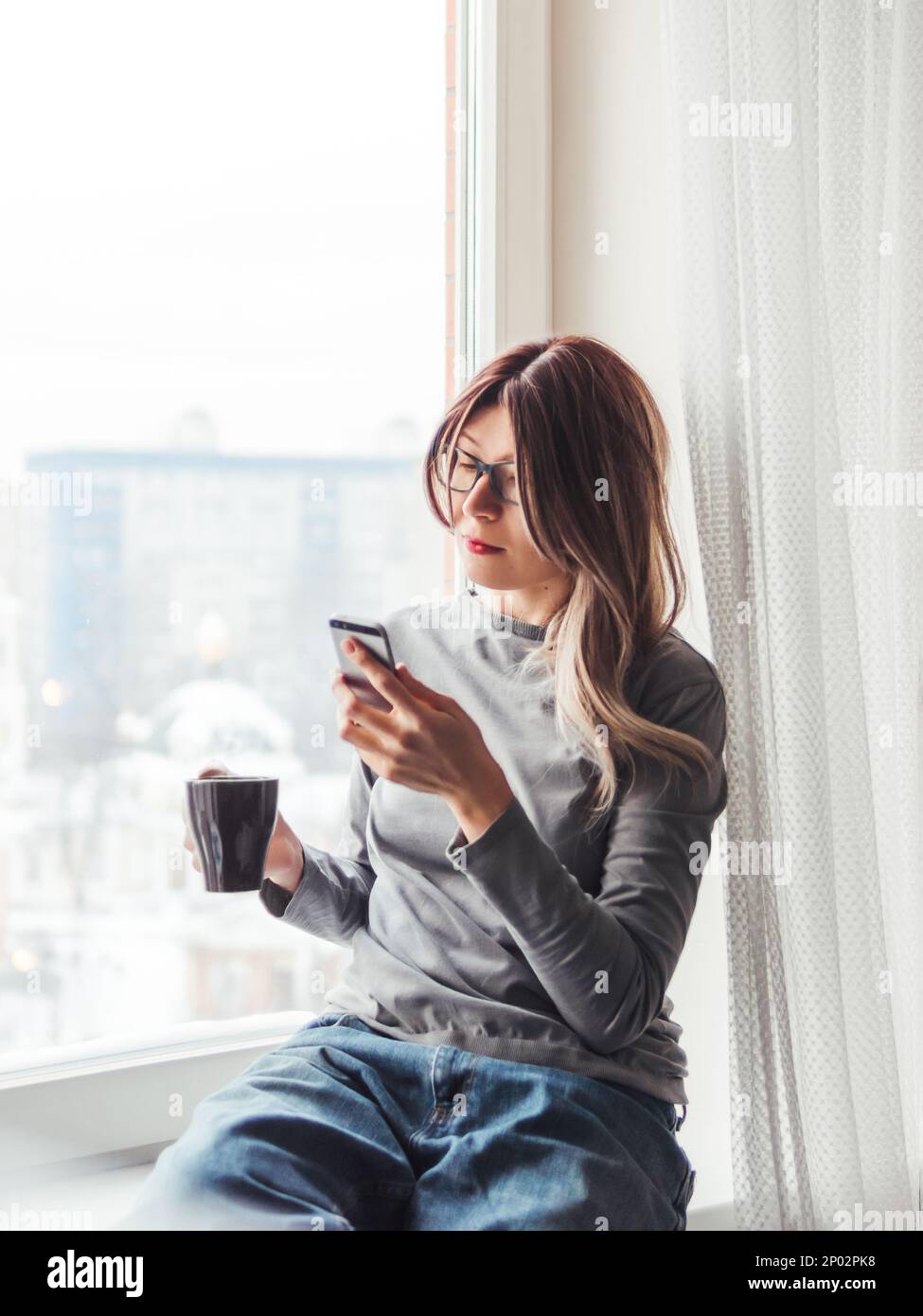 Une femme réfléchie avec des lunettes et une tasse de café chaud regarde son smartphone.Informations sur les médias en ligne. Banque D'Images