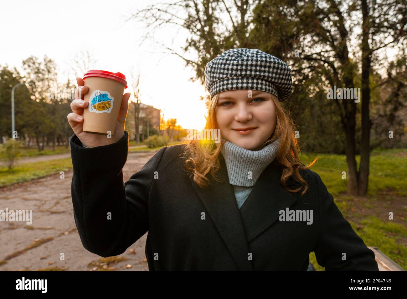 Ukraine. Soirée. Coucher de soleil. Parc de la ville. La fille tient dans ses mains une tasse de café avec un autocollant patriotique "tout sera l'Ukraine". Promenades en plein air Banque D'Images