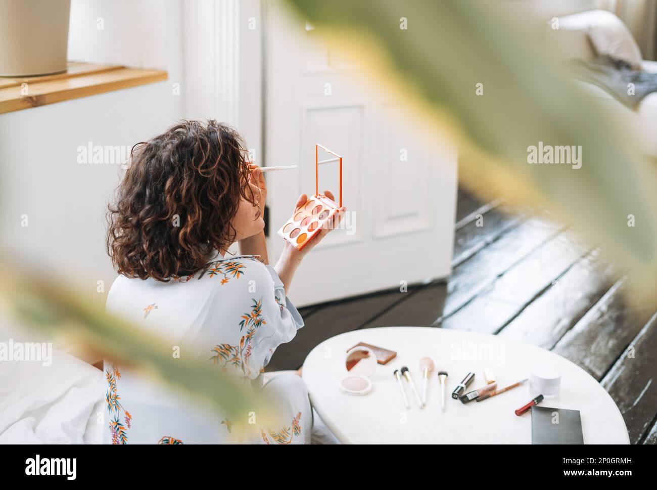 Jeune femme brune en pyjama bleu applique le maquillage avec le visage sculptant palette dans les mains à la maison Banque D'Images