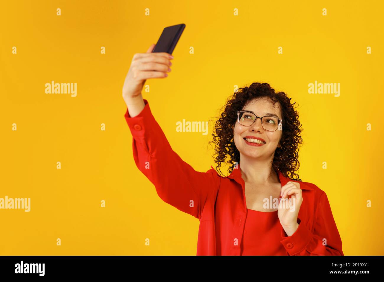 Jolie fille en forme de curly avec des lunettes prenant un selfie. Une jeune femme prend une photo d'elle-même au téléphone. Studio portrait de femme sur un fond jaune vif Banque D'Images