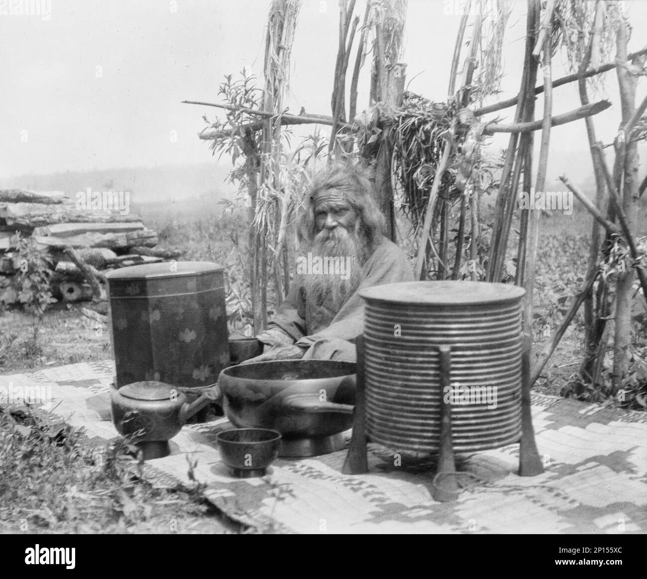 Ainu homme assis à l'extérieur sur un tapis recouvert de récipients en argile, 1908. Banque D'Images