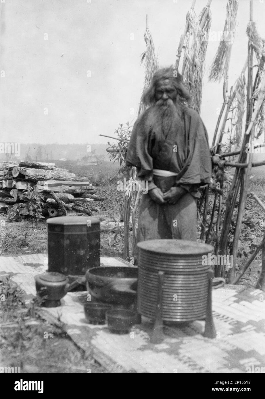 Ainu homme debout à l'extérieur sur un tapis recouvert de récipients en argile, 1908. Banque D'Images